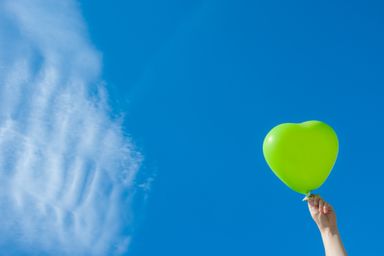 Hand holding a green heart-shaped balloon against a blue sky.